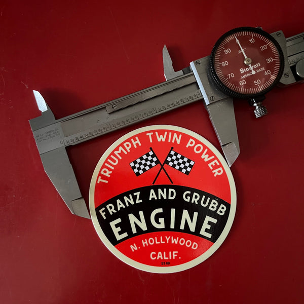 A round vintage-style red, black, and cream engine decal reading 'Triumph Twin Power Franz and Grubb Engine' is shown on a red background with a metal caliper measuring its diameter. The caliper brand 'Starrett' is visible.
