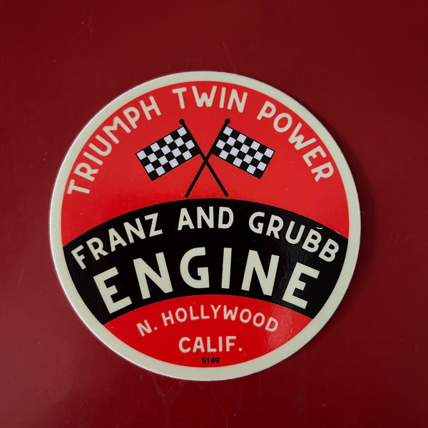 A circular vintage-style Triumph Twin Power engine sticker or decal reading 'Franz and Grubb Engine' with crossed checkered flags on a red background, shown against a darker red backdrop.